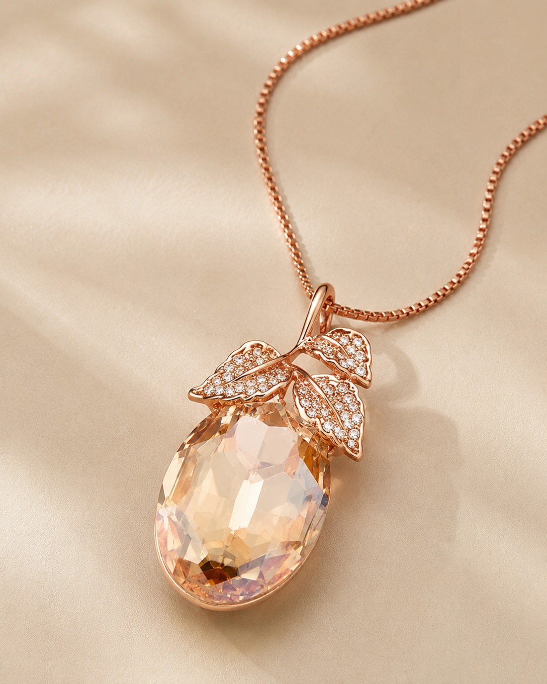 Rose gold pendant necklace featuring a faceted peach-colored teardrop gemstone pendant with leaf-shaped pavé accents, displayed on a soft beige satin background.
