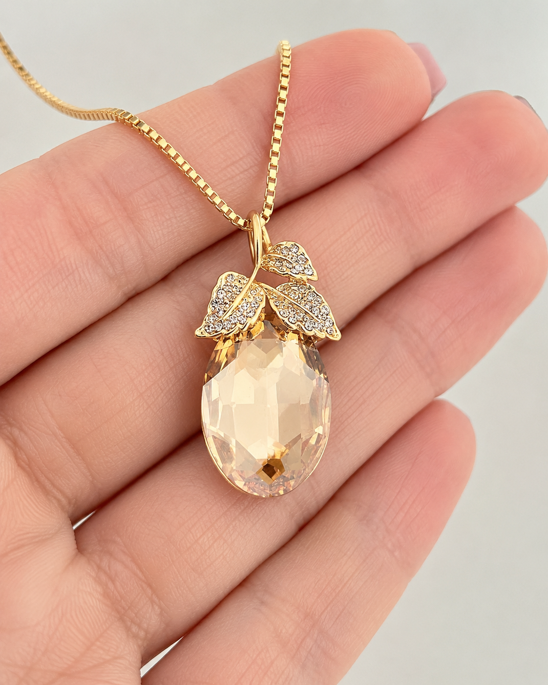 A gold pendant necklace with a faceted peach-colored teardrop gemstone and leaf-shaped settings, held in a person's hand against a light gray background.