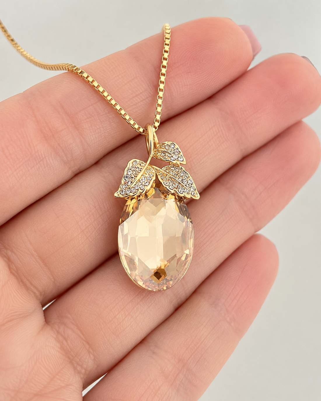 A gold pendant necklace with a faceted peach-colored teardrop gemstone and leaf-shaped settings, held in a person's hand against a light gray background.
