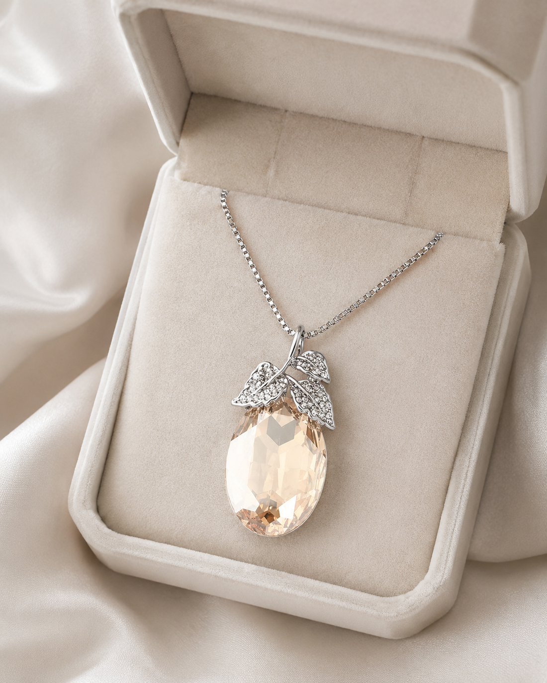 A silver pendant necklace with a large teardrop-shaped pale peach crystal and leaf-shaped crystal accents, displayed in an open cream-colored jewelry box on a soft satin fabric background.
