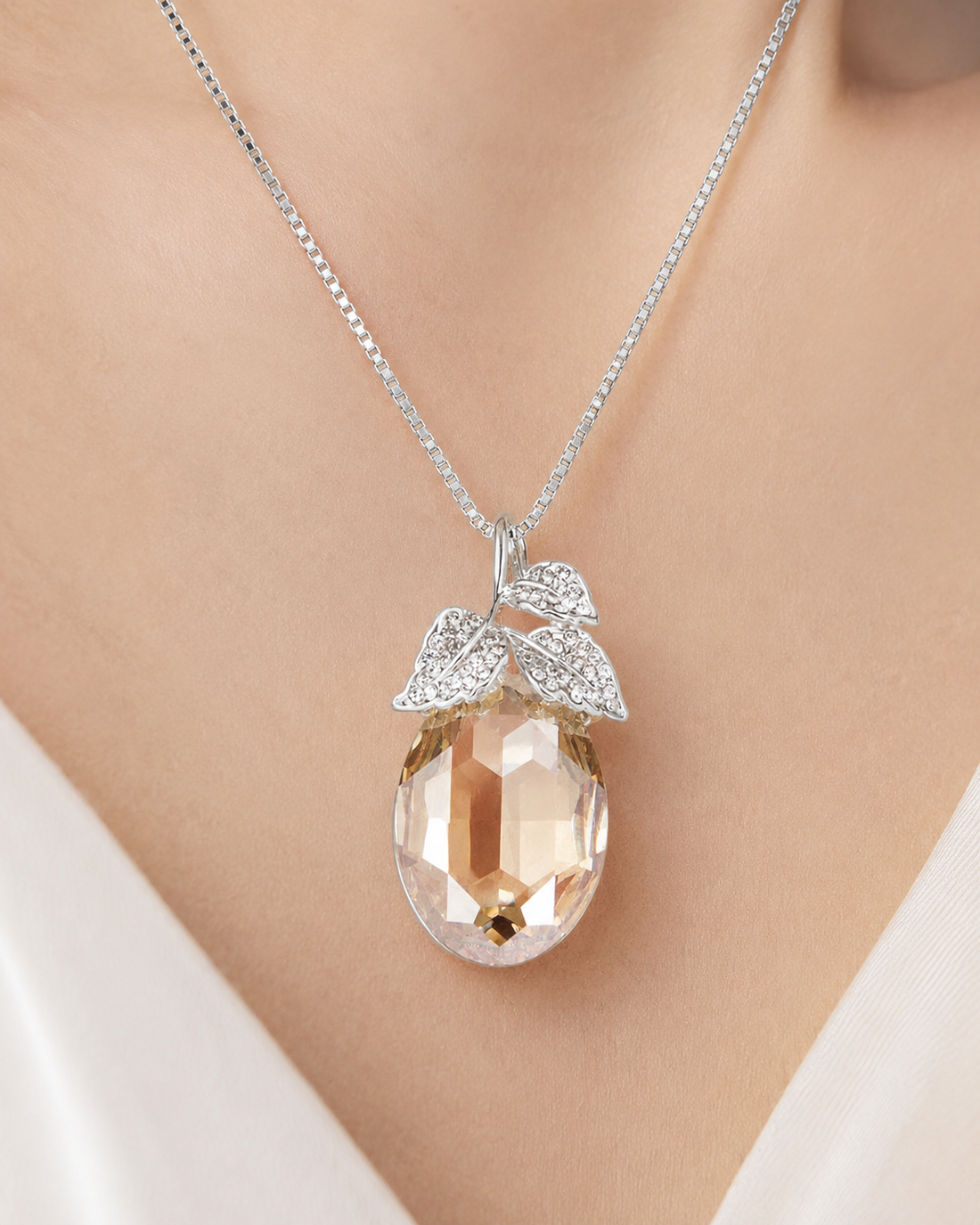 A silver pendant necklace with a faceted peach-colored teardrop gemstone and leaf-shaped crystal details, shown worn on a model's neck.
