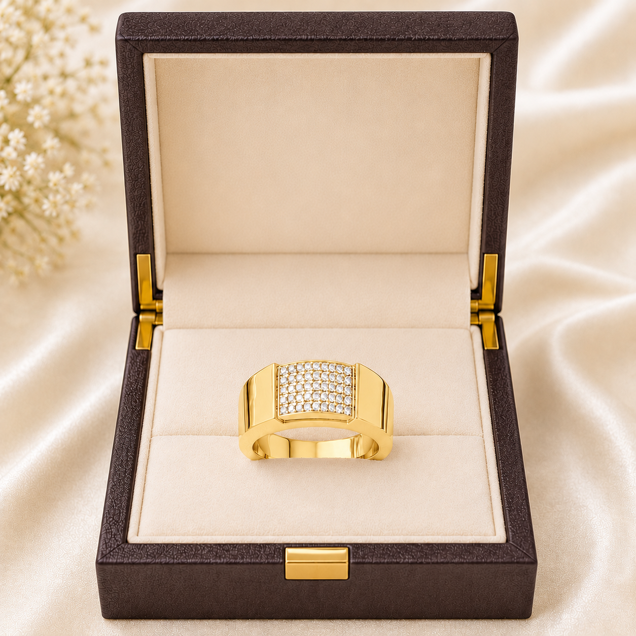 A gold ring with a square pavé crystal face sits in an open dark brown jewelry box with a cream velvet interior, set against a soft beige satin background with small white flowers in the corner.