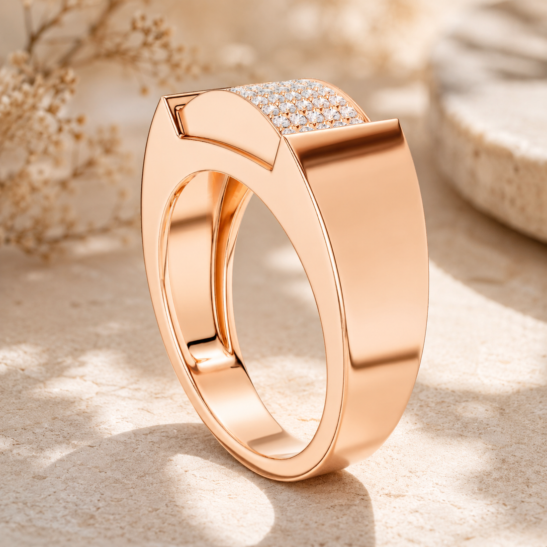 A polished rose gold ring with a pavé diamond top, displayed on a textured beige stone surface with soft, blurred dried flowers in the background.