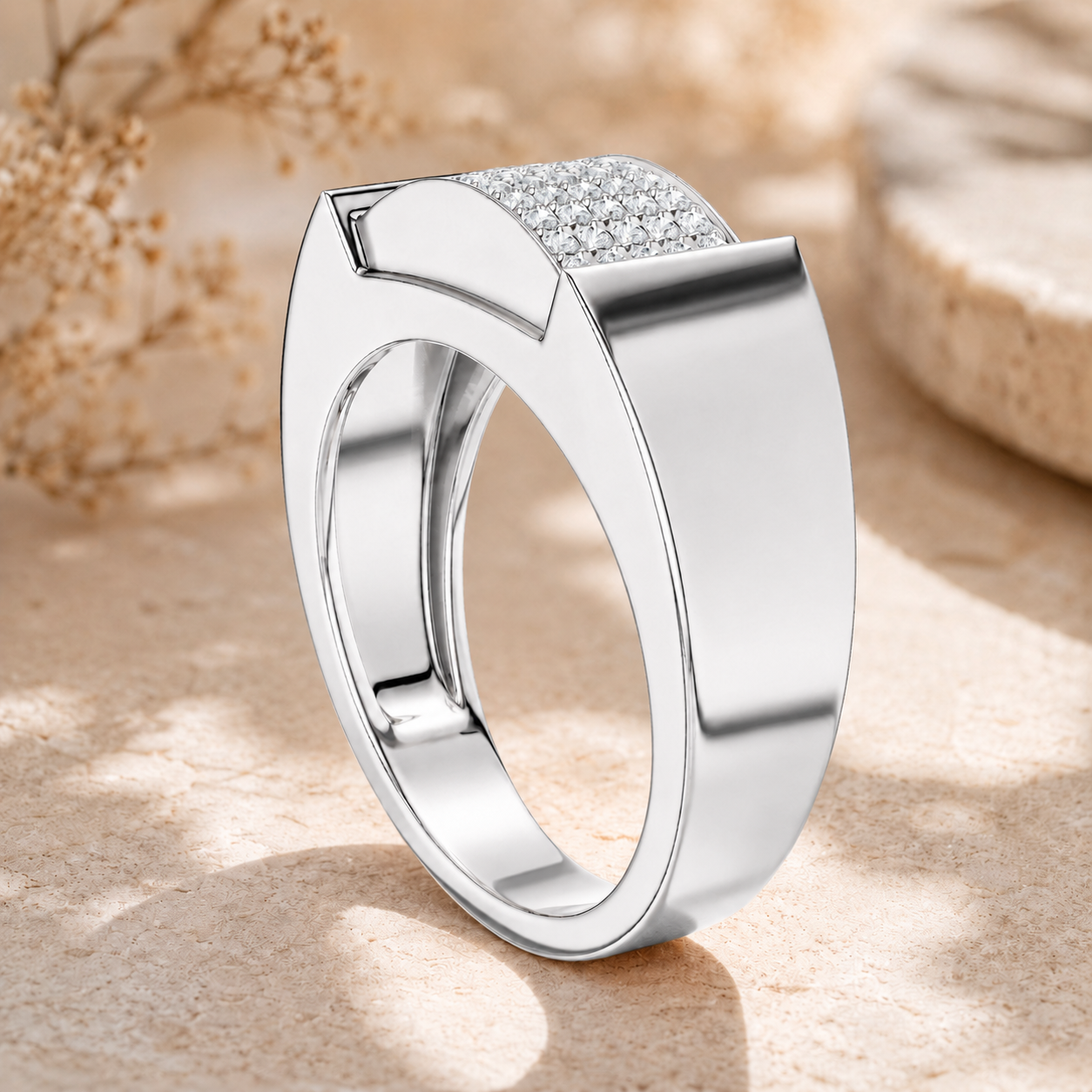 A polished silver-tone ring with a curved top featuring a row of clear pavé stones, displayed on a textured beige stone surface with soft blurred dried flowers in the background.