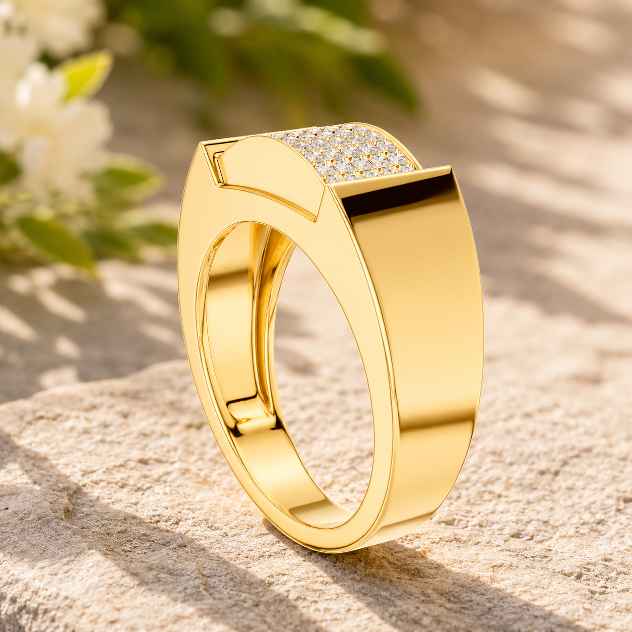 A polished yellow gold ring with a pavé diamond band, displayed on a textured stone surface with soft-focus white flowers and green leaves in the background.