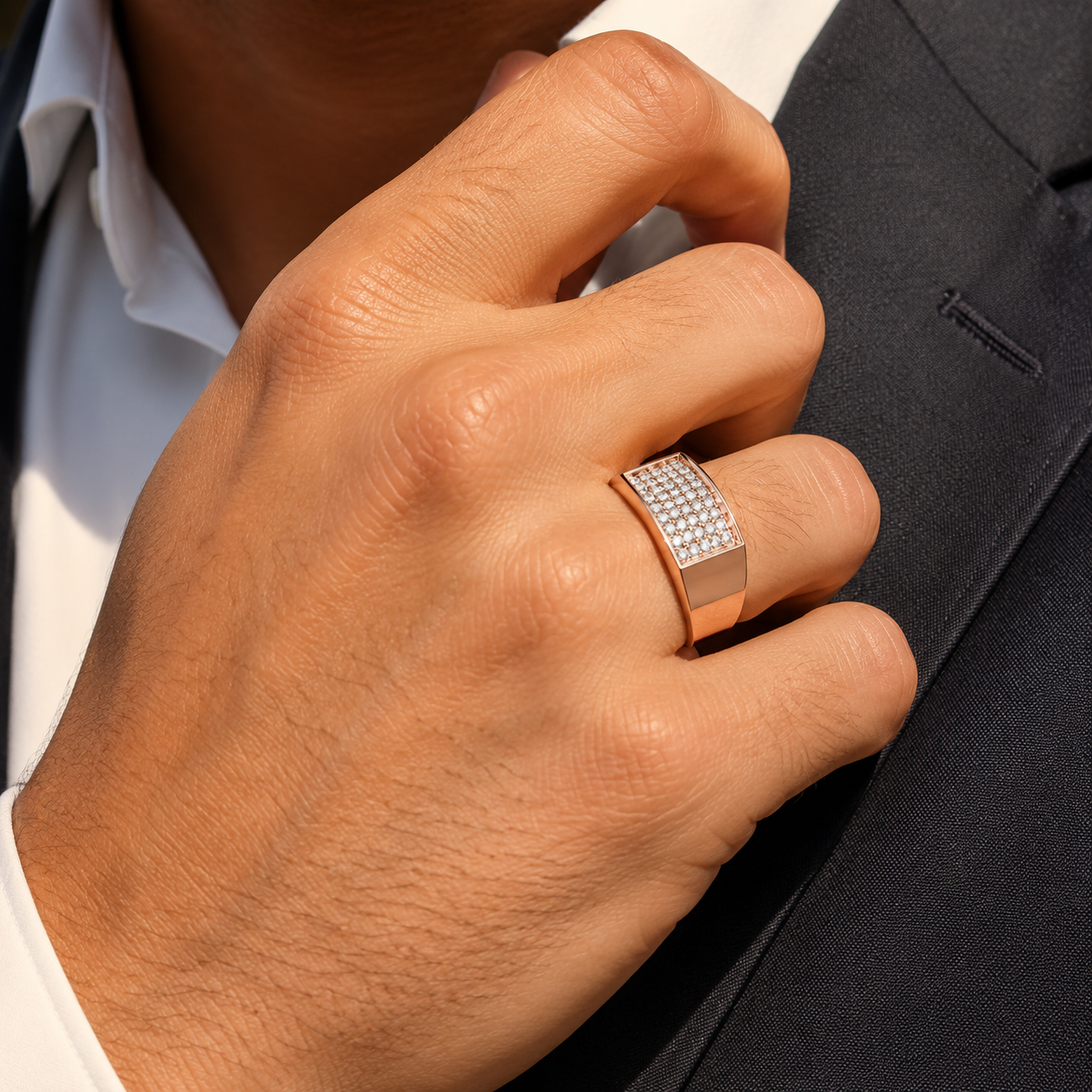 A close-up of a man's hand wearing a wide rose gold ring set with small clear stones, styled with a white collared shirt and dark gray suit jacket.