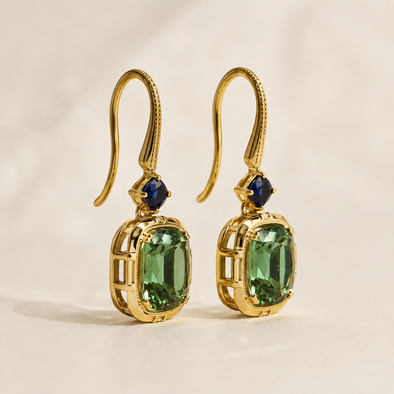 A pair of gold drop earrings with rectangular green gemstones and small dark blue accent stones, photographed on a clean light beige background.