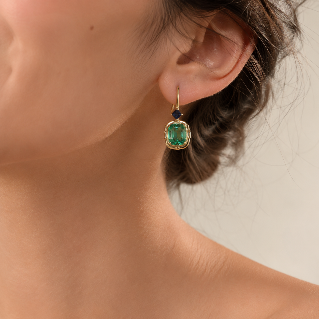 A close-up of a woman's ear wearing a gold drop earring with a cushion-cut green gemstone and a small dark blue accent stone above it.