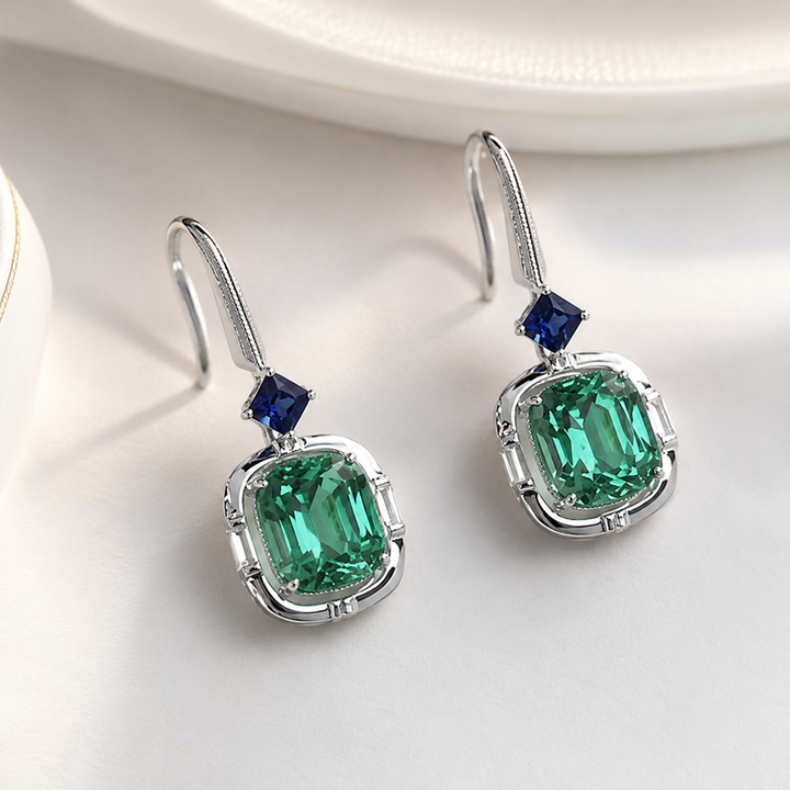 A pair of silver drop earrings featuring a large emerald-cut green center stone and a smaller dark blue accent stone above it, displayed on a clean white surface.