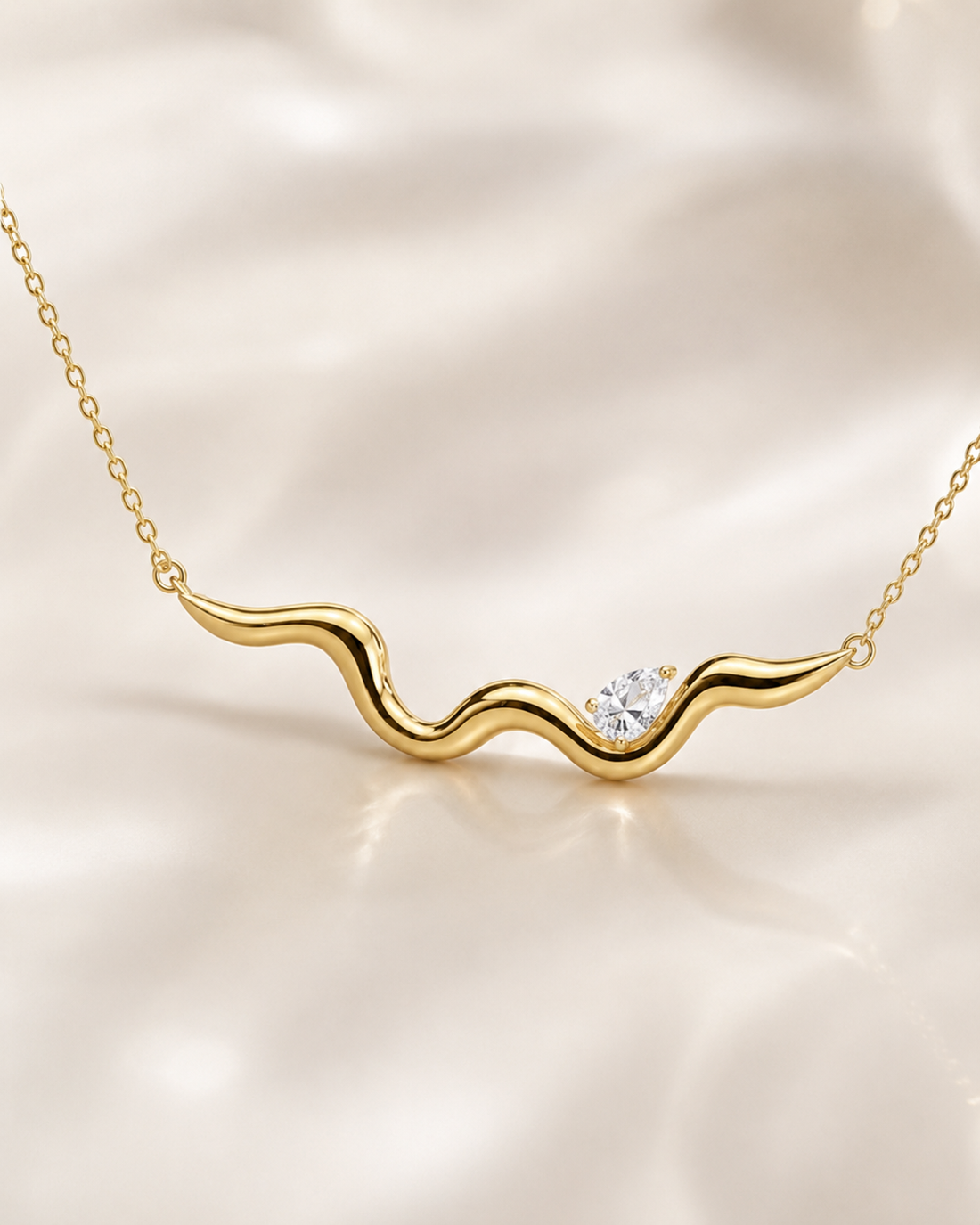 A gold wavy necklace with a small clear gemstone, displayed on a soft, draped cream satin fabric background.