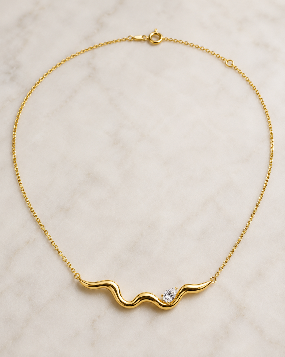 A delicate yellow gold chain necklace with a wavy, undulating bar pendant featuring a small clear gemstone, displayed on a light marble surface.