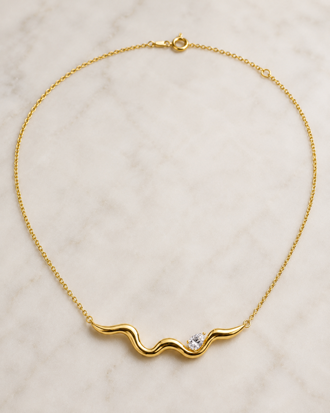 A delicate yellow gold chain necklace with a wavy, undulating bar pendant featuring a small clear gemstone, displayed on a light marble surface.