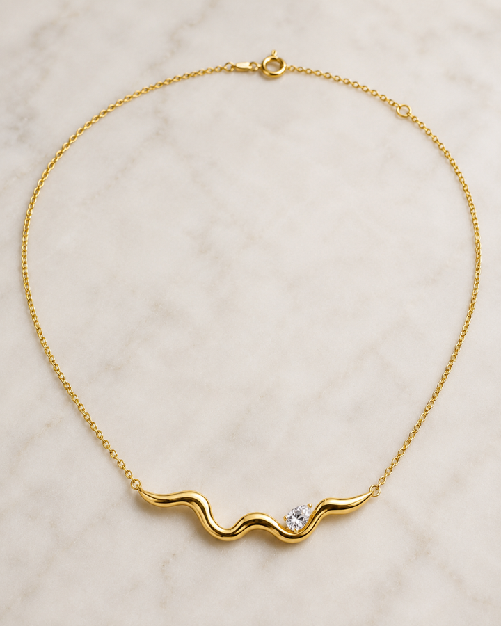 A delicate yellow gold chain necklace with a wavy, undulating bar pendant featuring a small clear gemstone, displayed on a light marble surface.