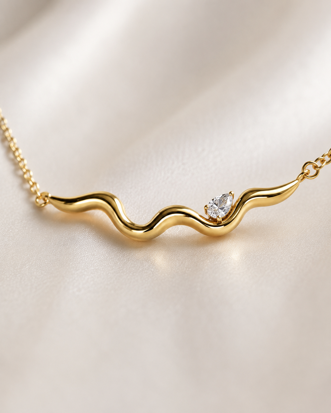 A gold wavy necklace with a small clear heart-shaped gemstone, displayed on a soft beige satin fabric background.