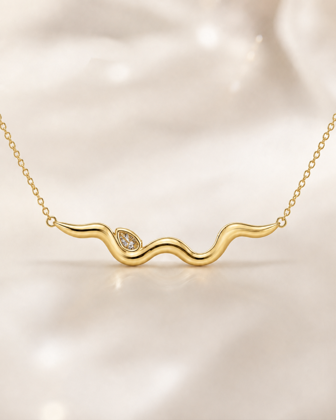 A gold wavy necklace with a small teardrop-shaped clear gemstone accent, displayed on a soft, light beige fabric background.