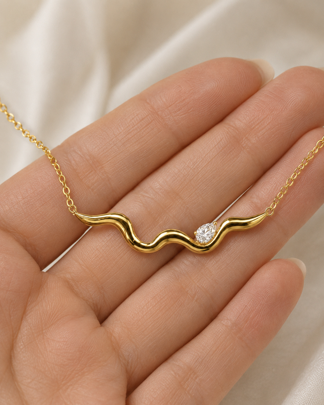 A gold wavy necklace with a small clear gemstone, held on a person's hand against a soft cream fabric background.
