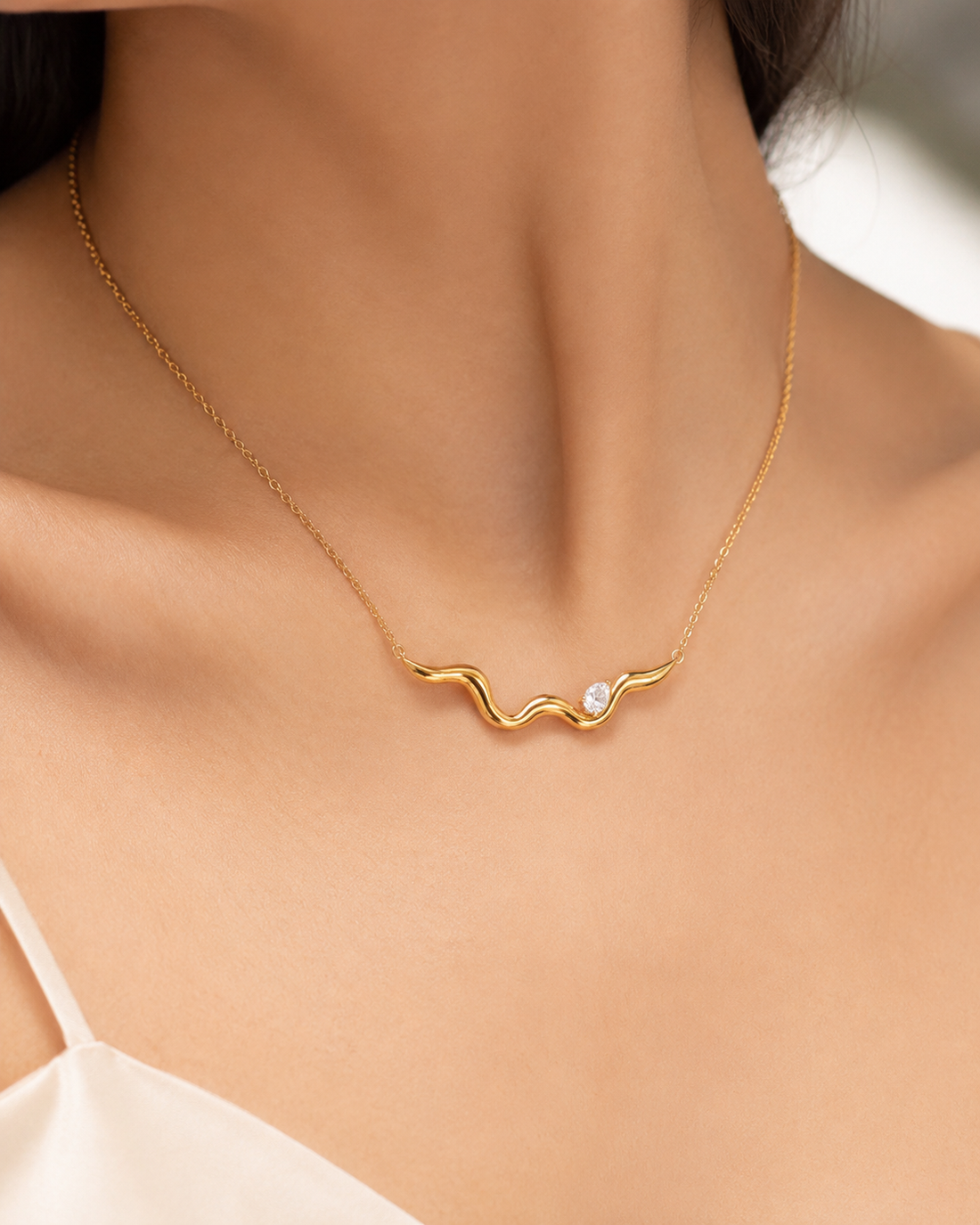A close-up of a model's neck and upper chest wearing a delicate gold chain necklace with a wavy, abstract pendant and a small clear stone.