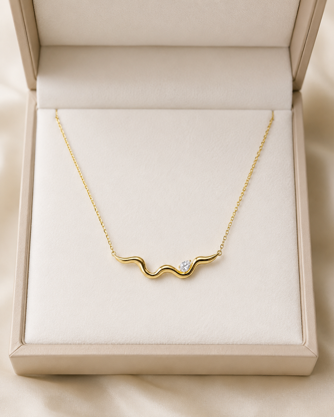 A gold wavy necklace with a small clear gemstone, presented in an open cream-colored jewelry box.