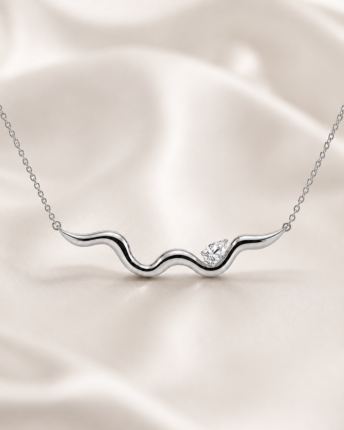 A silver wavy necklace with a small clear gemstone accent, displayed on a soft beige satin fabric background.