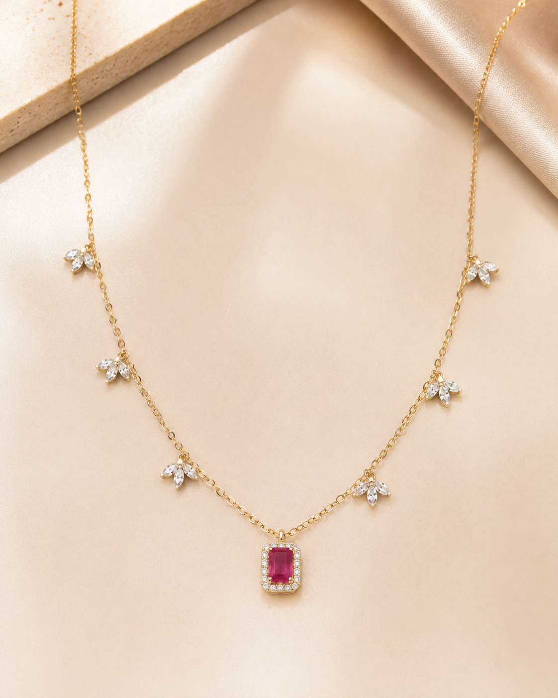 A delicate gold chain necklace featuring a central rectangular pink gemstone pendant surrounded by small clear stones, with five small star-shaped floral clusters spaced along the chain, displayed on a soft beige satin background.