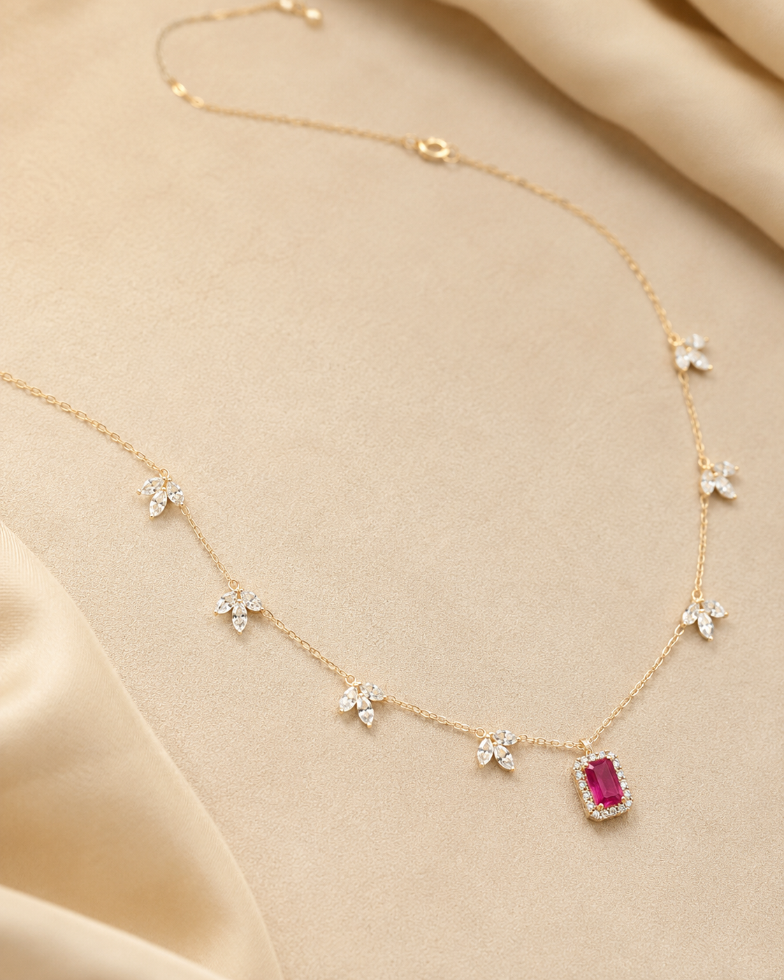 A delicate gold chain necklace with small white floral motifs and a central rectangular pink gemstone pendant, displayed on a soft beige satin fabric background.