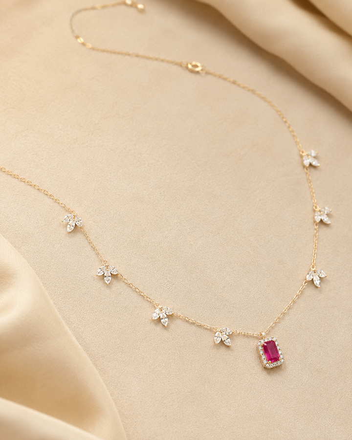 A delicate gold chain necklace with small white floral motifs and a central rectangular pink gemstone pendant, displayed on a soft beige satin fabric background.