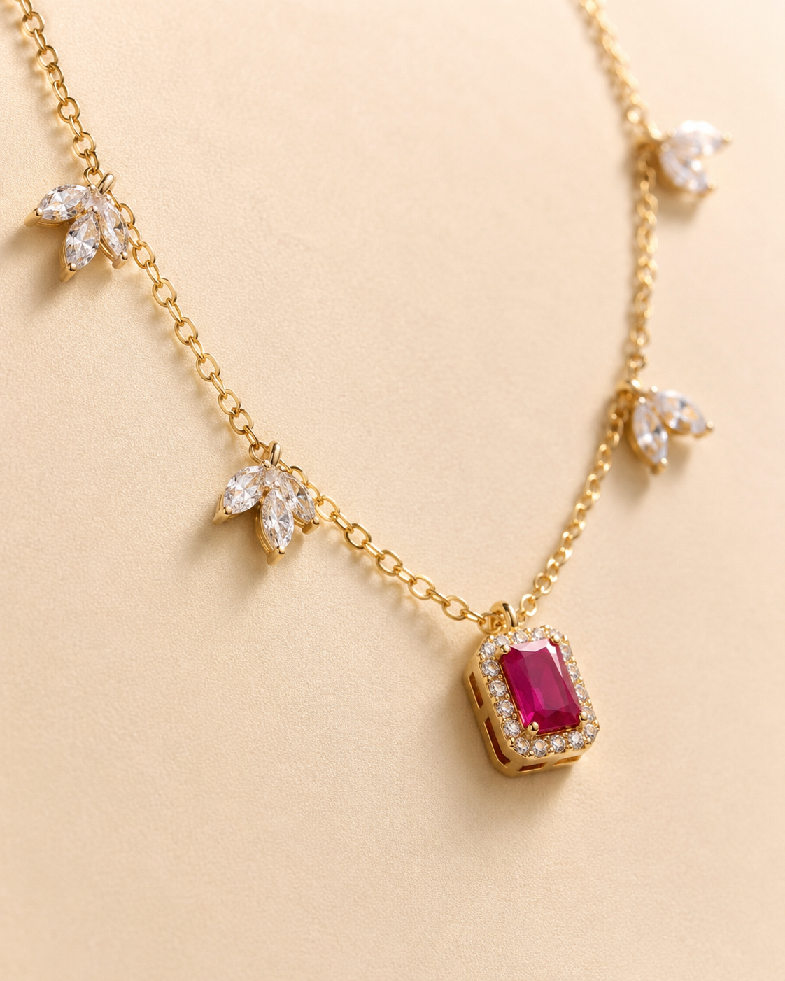 A gold chain necklace featuring a central rectangular ruby pendant surrounded by small clear stones, with five matching leaf-shaped clear stone clusters spaced along the chain, displayed on a warm beige background.