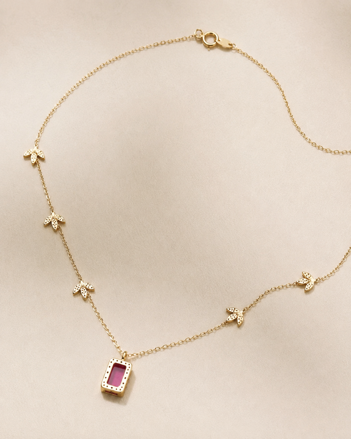 A delicate gold chain necklace featuring a central rectangular pendant with a pink gemstone surrounded by small clear stones, accented by four small gold flower motifs spaced along the chain, displayed on a neutral beige background.