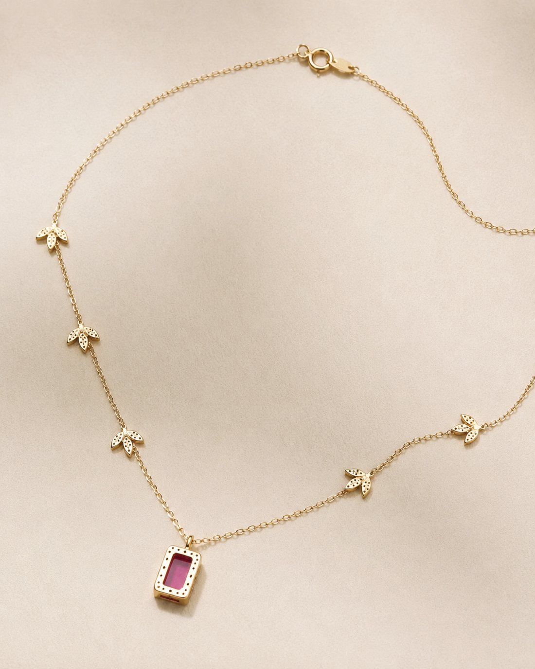 A delicate gold chain necklace featuring a central rectangular pendant with a pink gemstone surrounded by small clear stones, accented by four small gold flower motifs spaced along the chain, displayed on a neutral beige background.