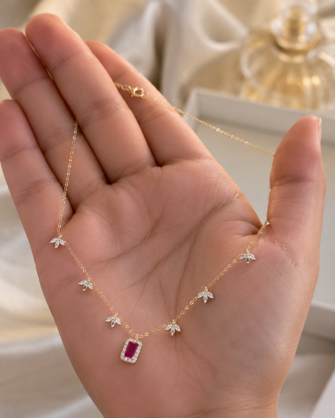 A delicate gold necklace with five small flower charms and a central rectangular pink gemstone pendant, displayed on an open hand.