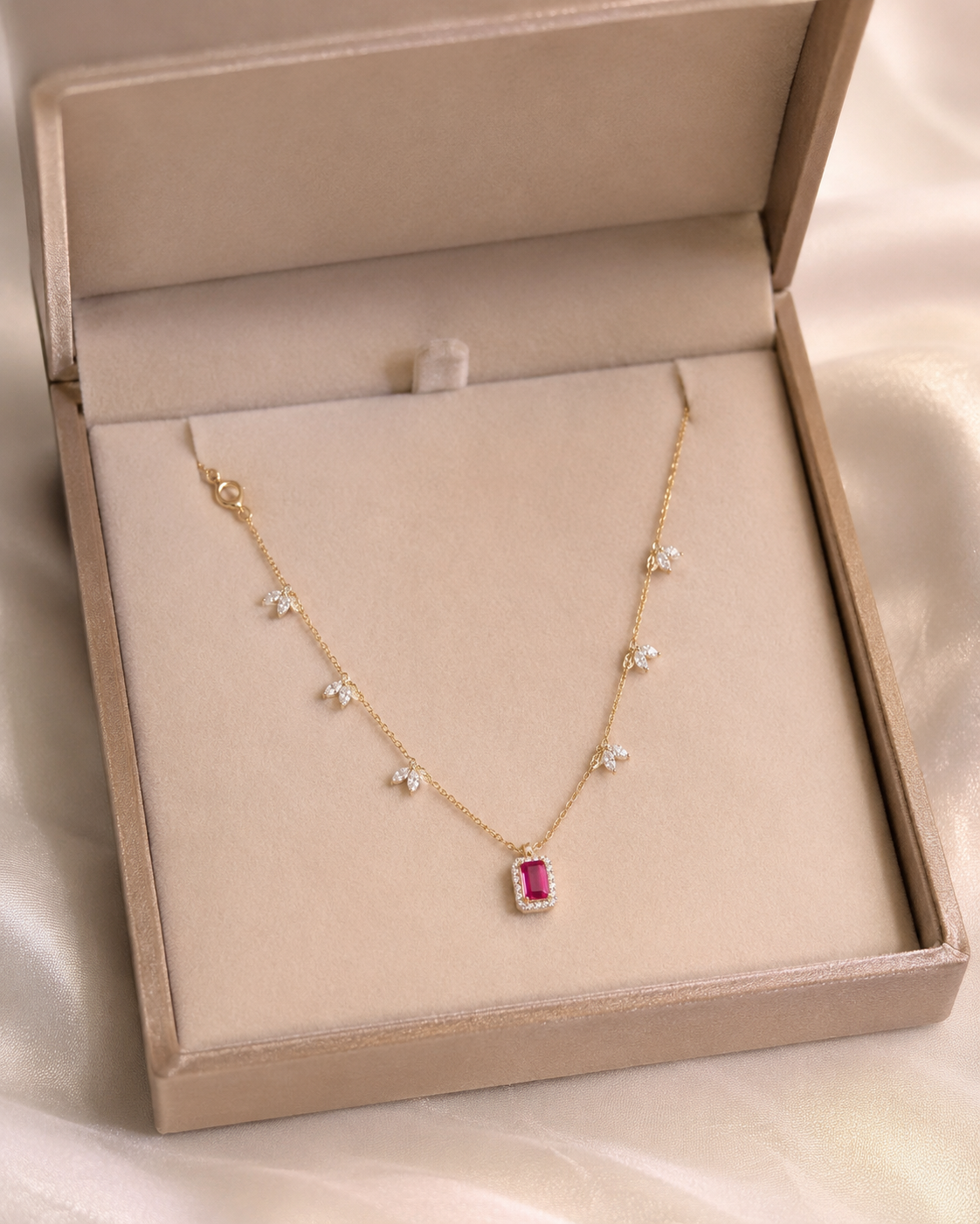 A delicate gold necklace with small star-shaped clear stones and a rectangular pink gemstone pendant, displayed inside a beige jewelry box on a soft satin fabric background.