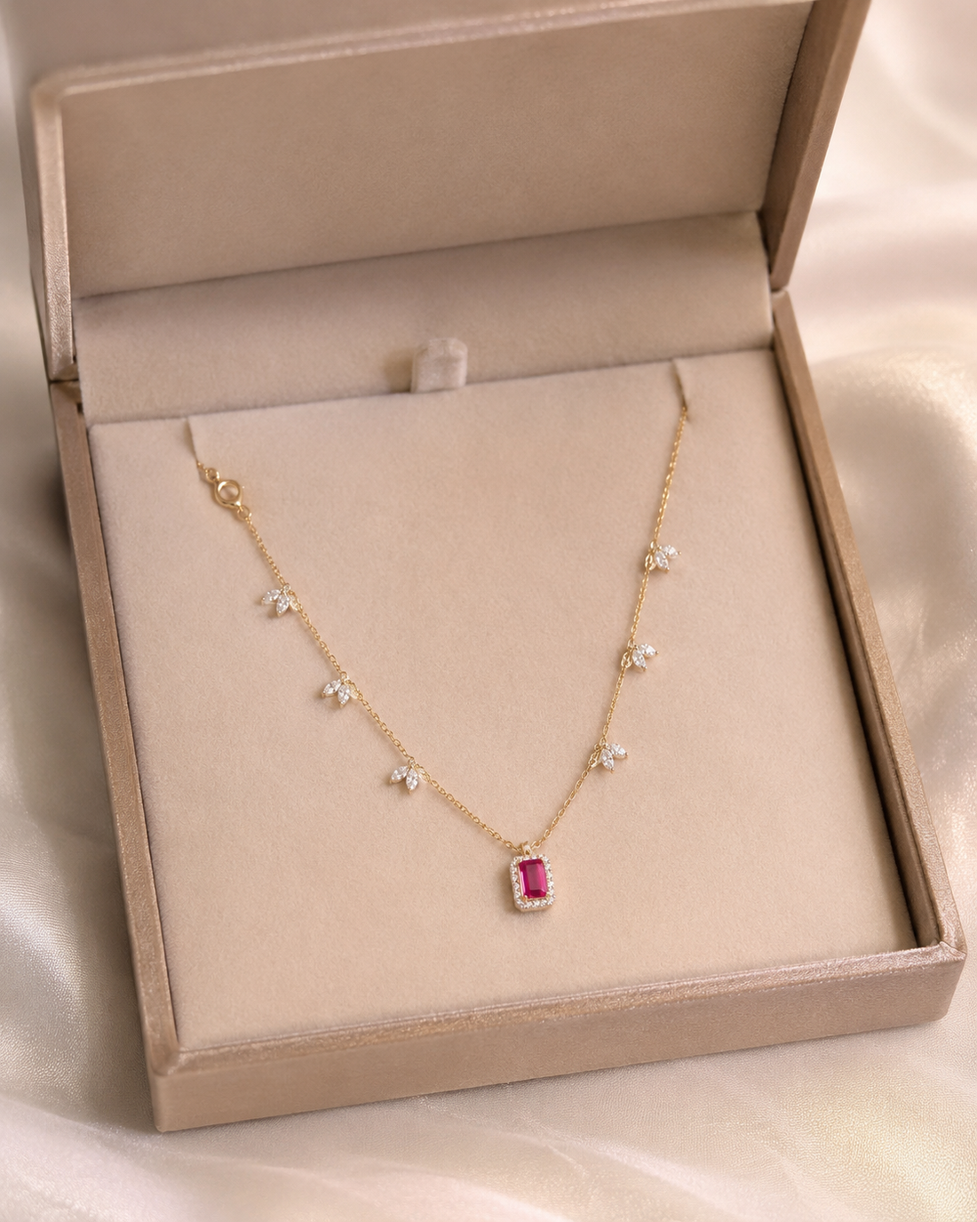 A delicate gold necklace with small star-shaped clear stones and a rectangular pink gemstone pendant, displayed inside a beige jewelry box on a soft satin fabric background.