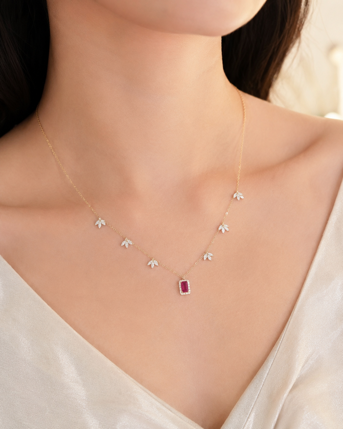 A delicate gold necklace worn by a model, featuring small white crystal-like flower charms on each side of a central rectangular pendant with a deep red gemstone.