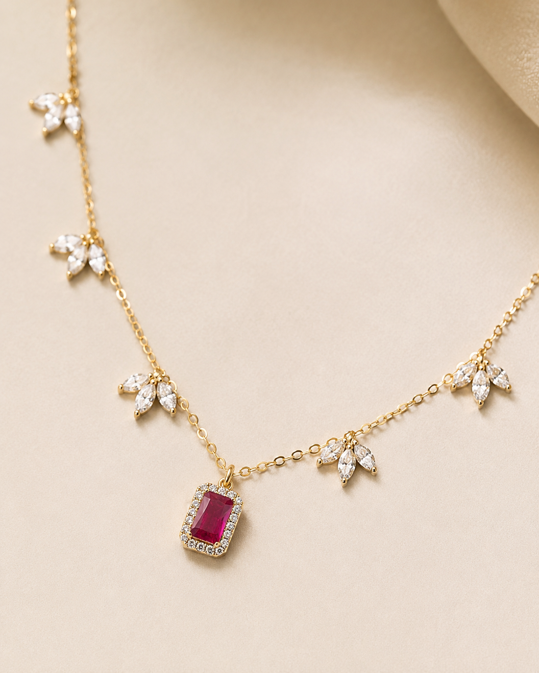 A gold chain necklace featuring a central rectangular pinkish-red gemstone pendant surrounded by small clear crystals, with additional crystal leaf motifs along the chain, displayed on a beige surface.