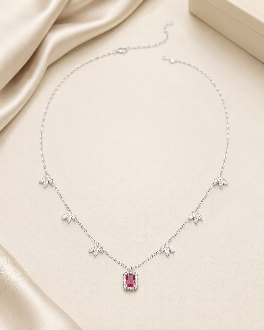 A delicate silver-tone necklace with a central rectangular pink gemstone pendant, accented by small butterfly motifs along the chain, displayed on a beige satin fabric background.