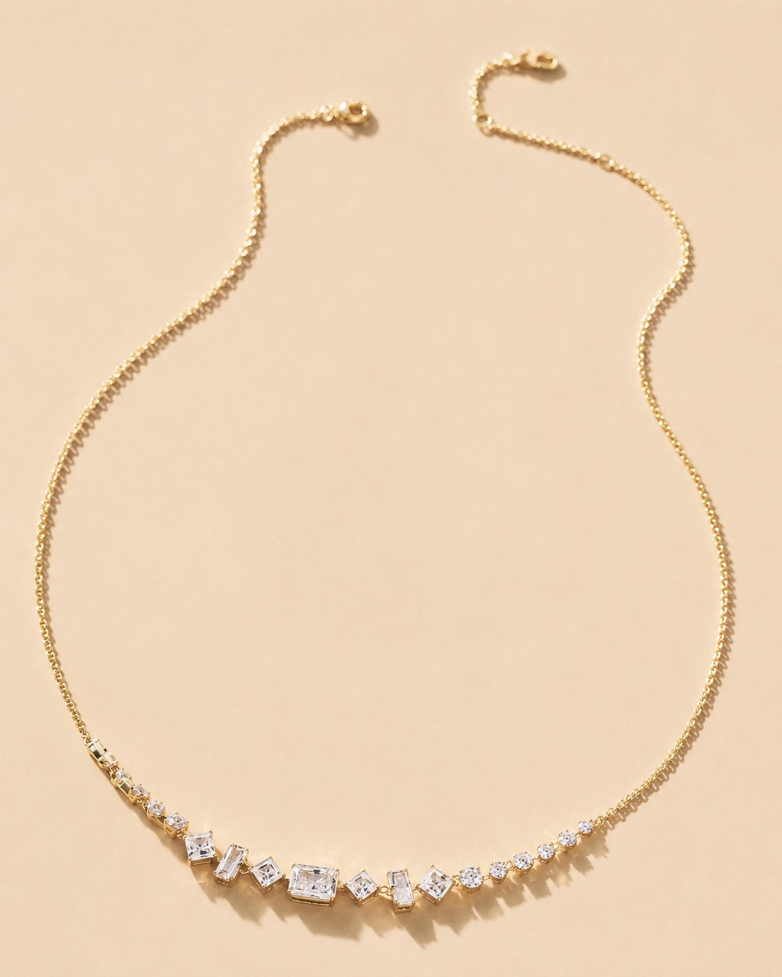 A delicate gold chain necklace with a curved bar of clear baguette and round-cut stones, displayed on a solid light beige background.