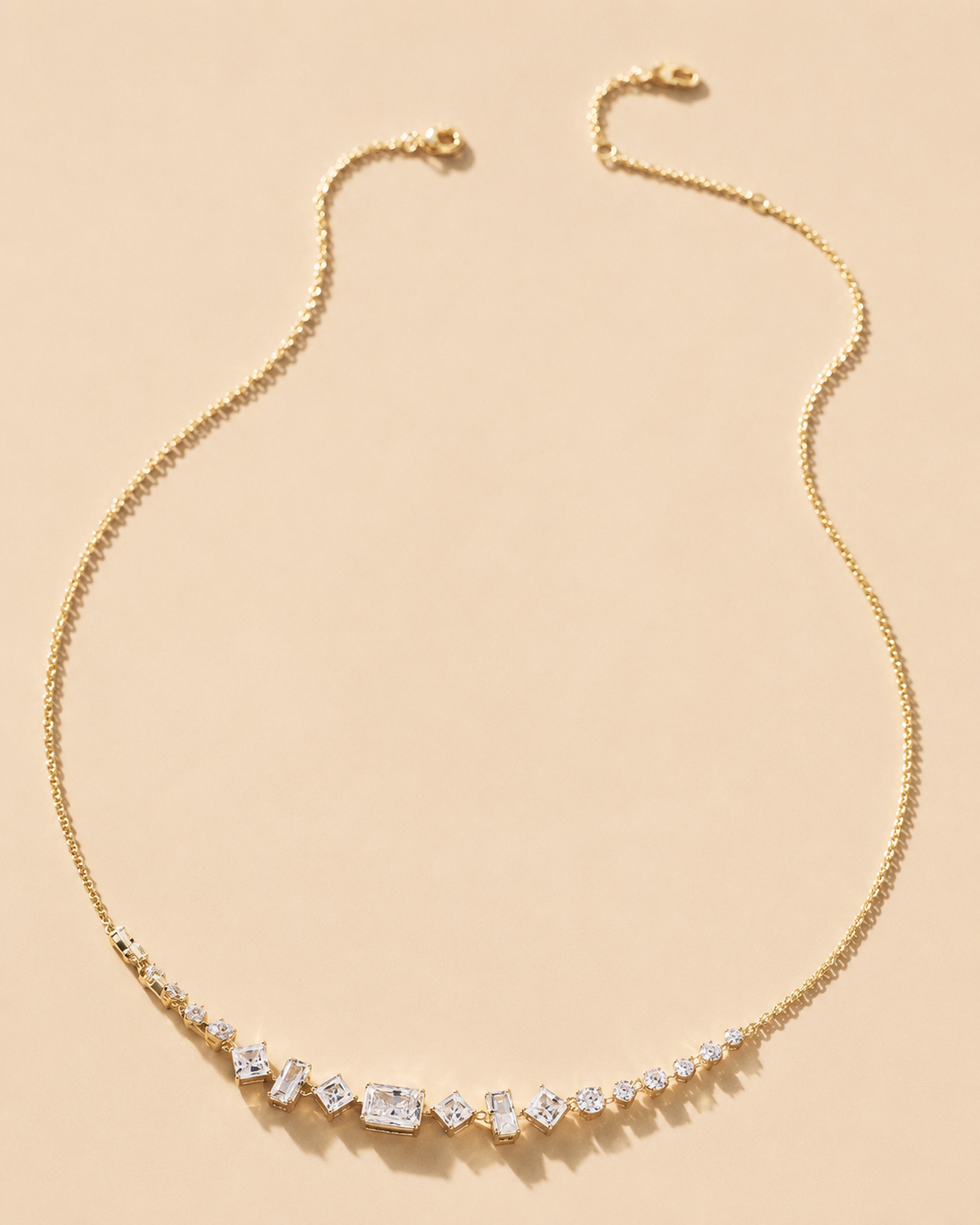 A delicate gold chain necklace with a curved bar of clear baguette and round-cut stones, displayed on a solid light beige background.