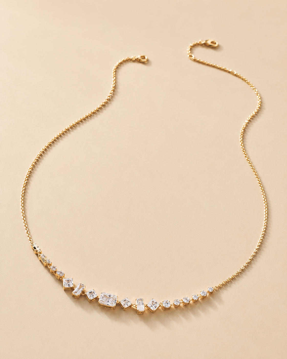 A delicate gold chain necklace featuring a curved row of clear heart-shaped and oval gemstones, displayed on a soft beige background.