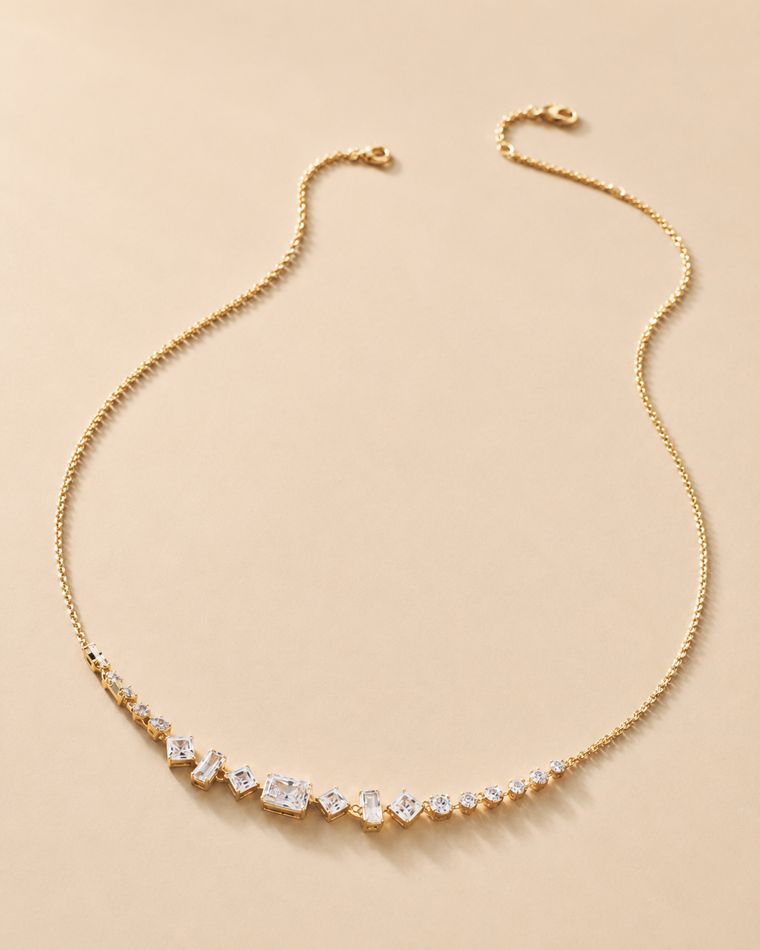 A delicate gold chain necklace featuring a curved row of clear heart-shaped and oval gemstones, displayed on a soft beige background.