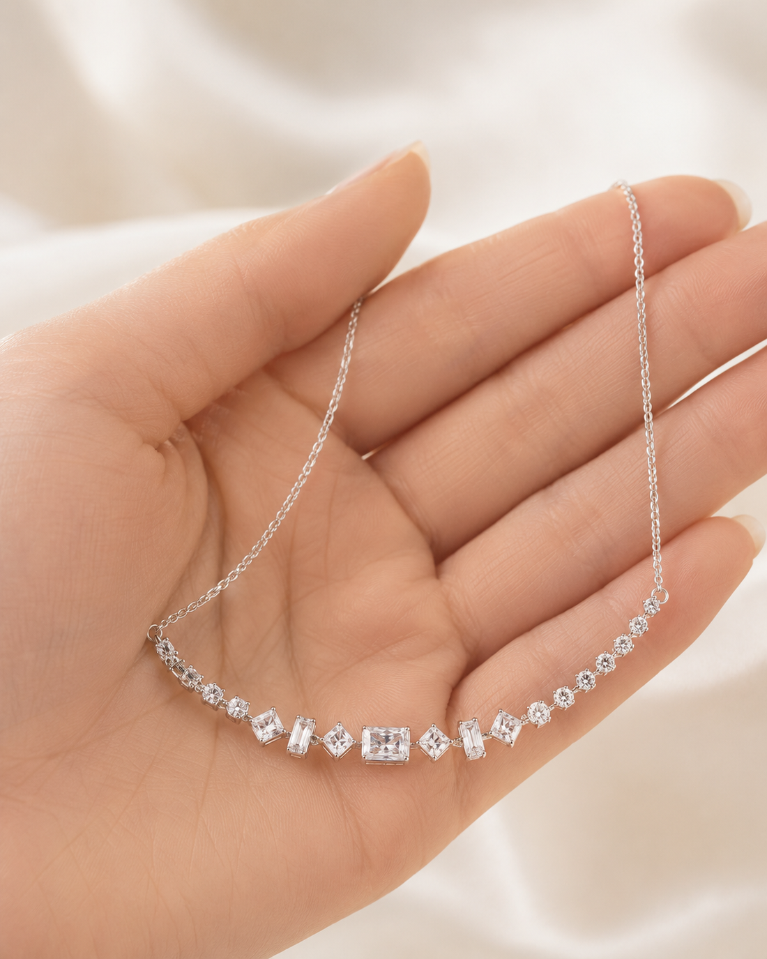 A delicate silver necklace with a row of clear gemstones displayed on an open hand against a soft white fabric background.