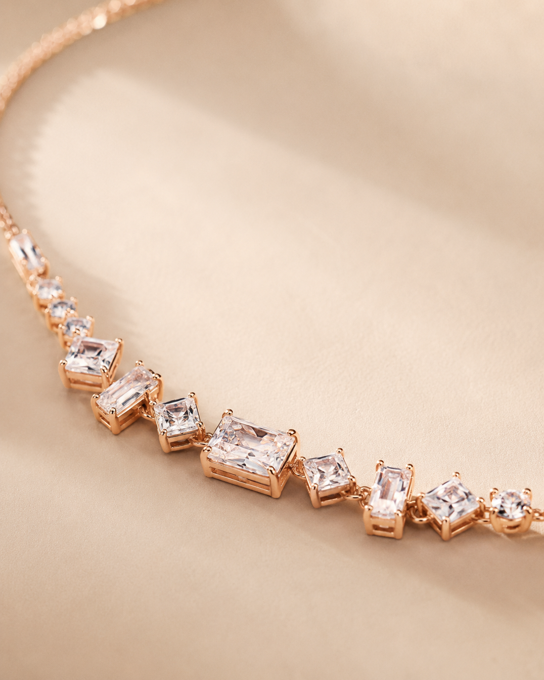 A close-up of a rose gold necklace featuring a series of clear rectangular and square gemstones in a graduated setting, displayed on a soft beige background.