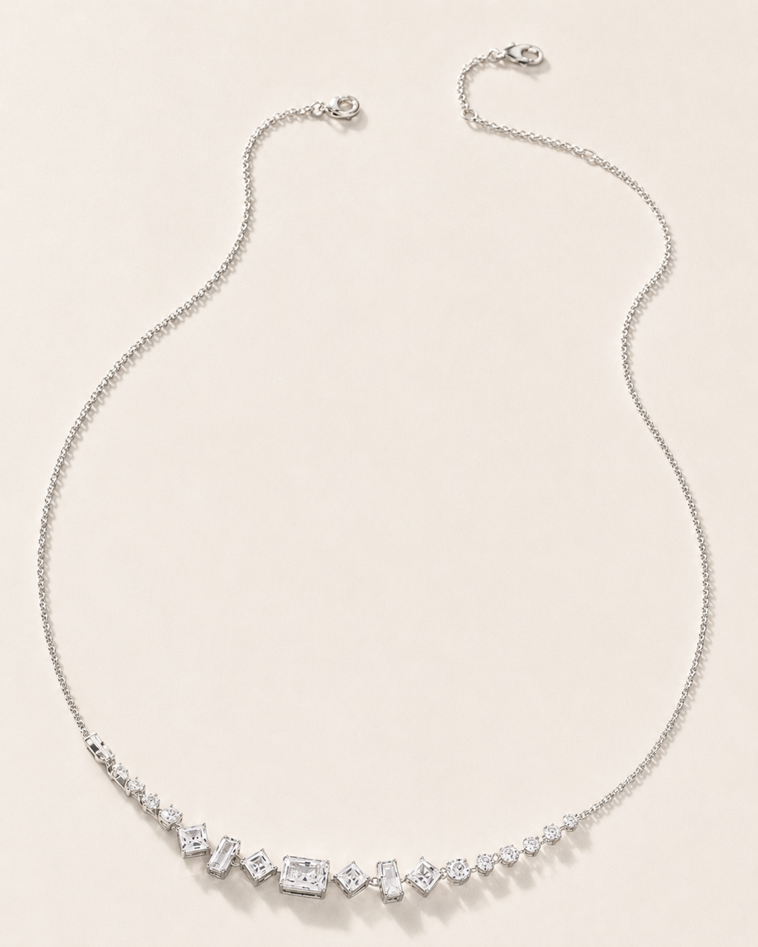 A delicate silver chain necklace featuring a curved bar of clear baguette and round-cut stones at the center, displayed on a light beige background.