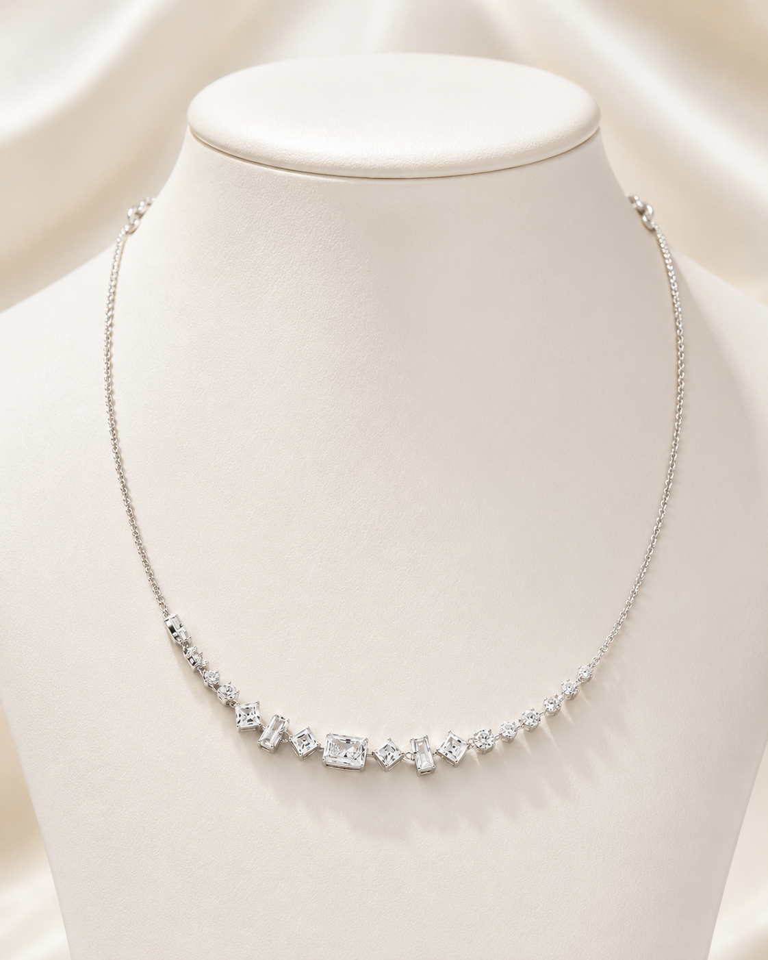 A delicate silver necklace with a row of clear baguette and round-cut stones, displayed on a white jewelry bust against a soft cream satin background.