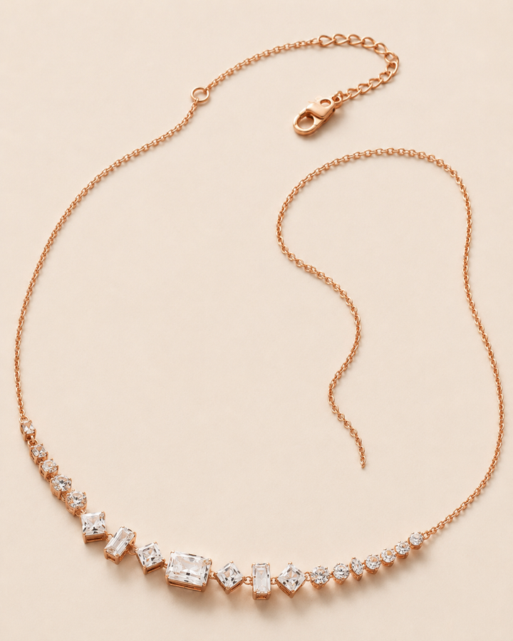 A rose gold necklace featuring a delicate chain and a curved row of clear baguette and round-cut gemstones, displayed on a light beige background.
