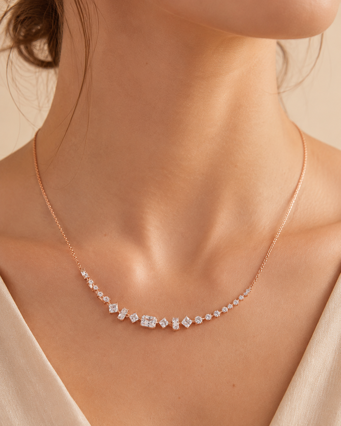 A close-up of a model's neck and upper chest wearing a delicate rose gold necklace with a curved row of clear gemstones.