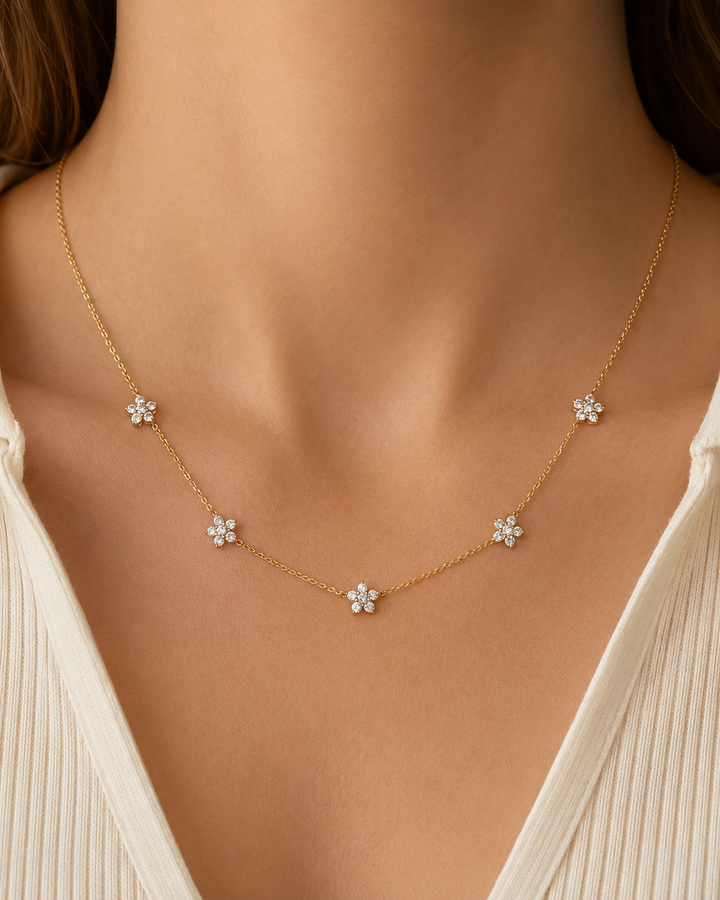 A delicate gold chain necklace with five small flower-shaped charms set with clear stones, worn on a model's neck over a cream-colored top.