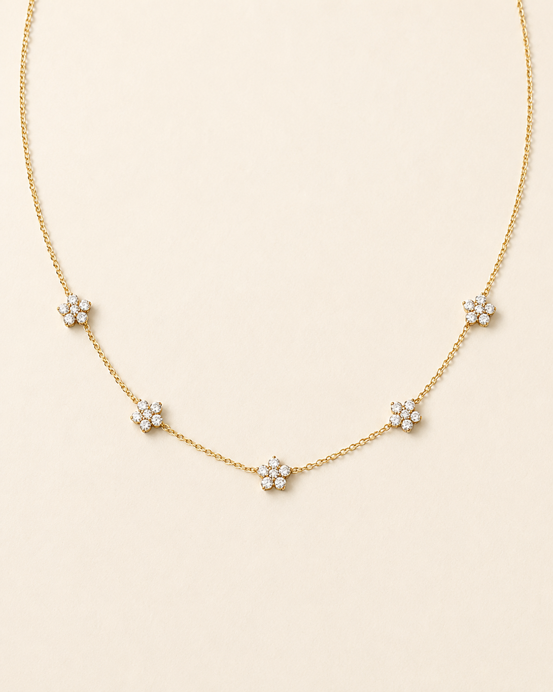 A delicate gold chain necklace featuring five small flower clusters, each set with clear round stones, arranged in a gentle arc on a light cream background.