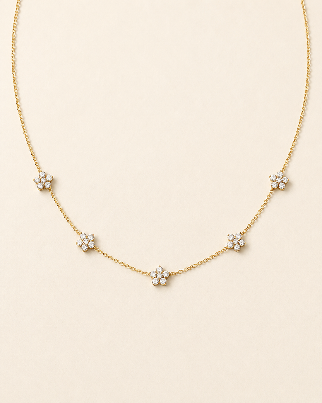 A delicate gold chain necklace featuring five small flower clusters, each set with clear round stones, arranged in a gentle arc on a light cream background.