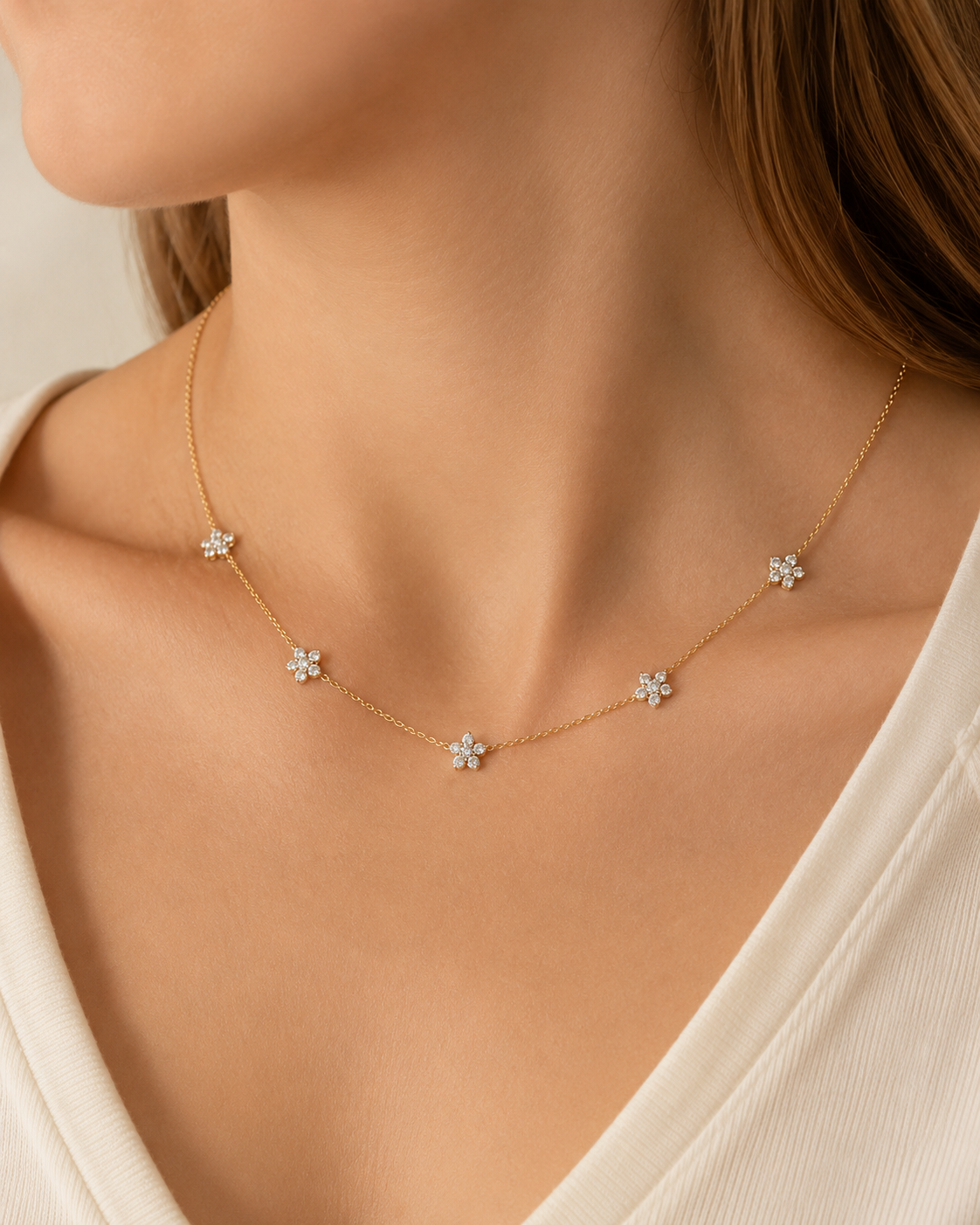 A delicate gold necklace with five small flower-shaped charms set with clear stones, worn on a model's neck.