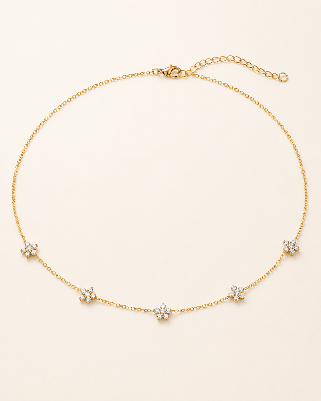 A delicate gold chain necklace featuring five small flower-shaped settings with clear stones, arranged in a circle on a light cream background.